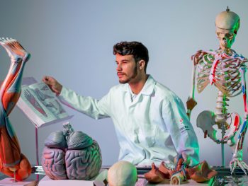 Estudante de Medicina do UNICEPLAC em laboratório de anatomia, analisando atlas anatômico ao lado de modelos de esqueleto humano, cérebro e músculos para estudo prático