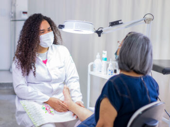 Mulher com jaleco de esteticista e máscara massageando os pés de uma mulher vestida de azul