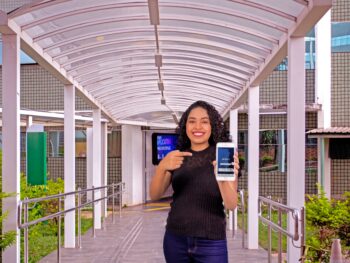 Imagme de uma mulher usando camiseta preta parada em frente a uma passarela com um celular na mão para ilustrar matéria sobre como usar a nota do Enem