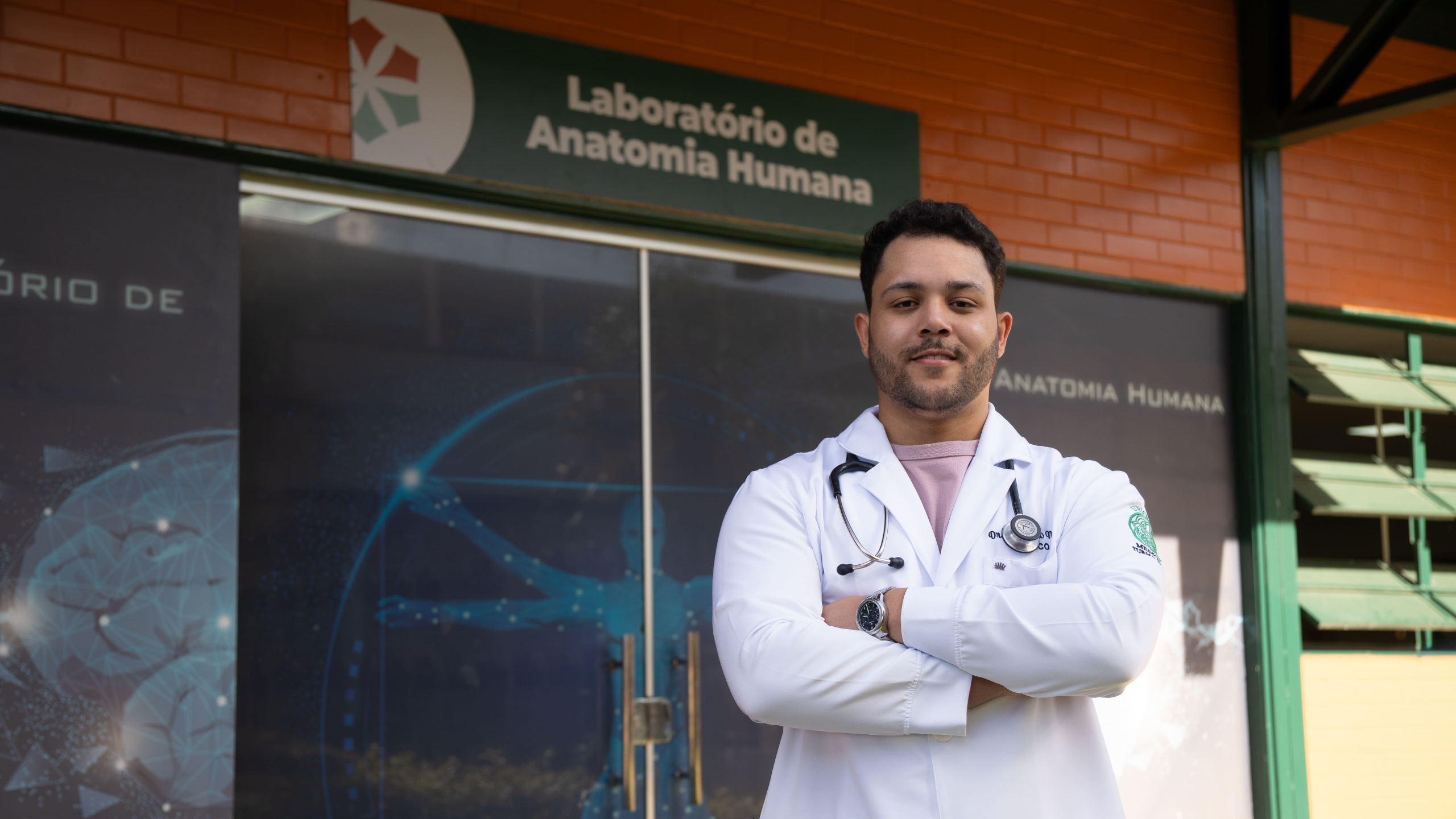 Imagem de um estudante do curso de Medicina do UNICEPLAC de braços cruzados e jaleco em frente ao Laboratório de Anatomia Humana